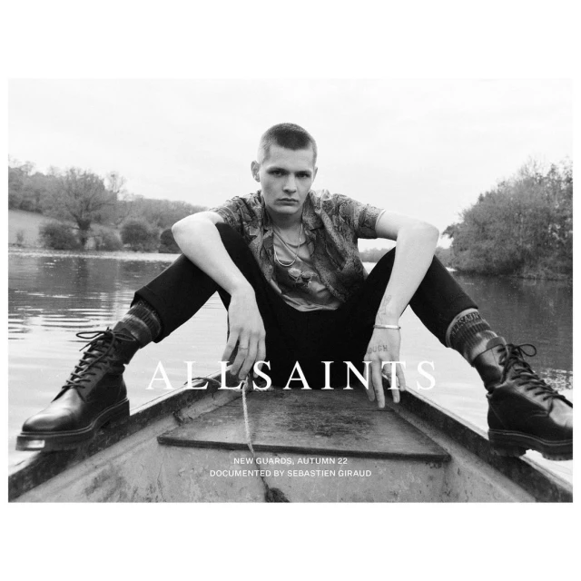 Monochrome portrait of a stylish adult male sitting in a boat on a river wearing boots, a patterned shirt, and dark pants in an outdoor setting.