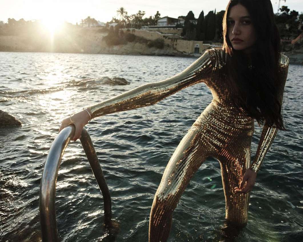 Woman in gold sequin jumpsuit standing waist-deep in ocean, holding onto metal ladder, with sun setting in background.