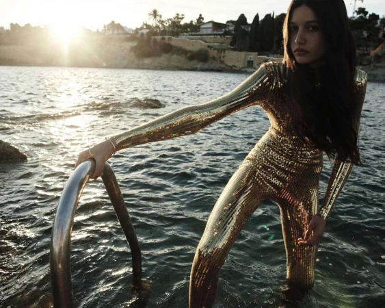 Woman in gold sequin jumpsuit standing waist-deep in ocean, holding onto metal ladder, with sun setting in background.