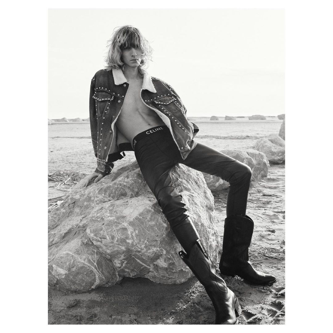A model with long hair poses on rocks in black jeans, cowboy boots and studded denim jacket in a black and white fashion shot.