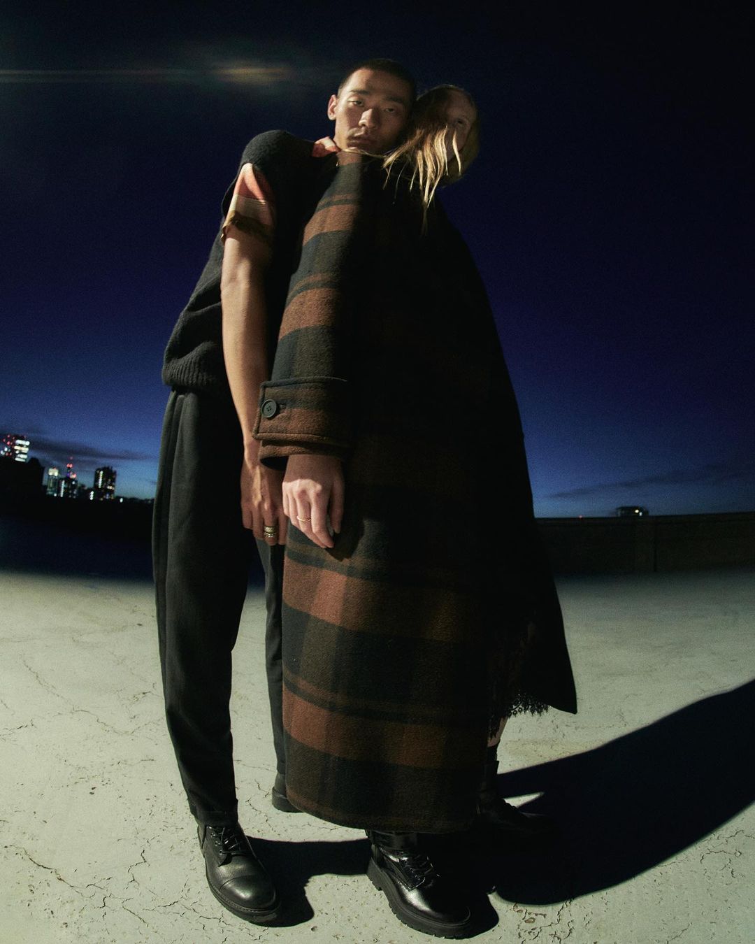 Two fashion models, one Asian man in a black sweater and one white woman in a coat, pose together on a rooftop in an urban environment.