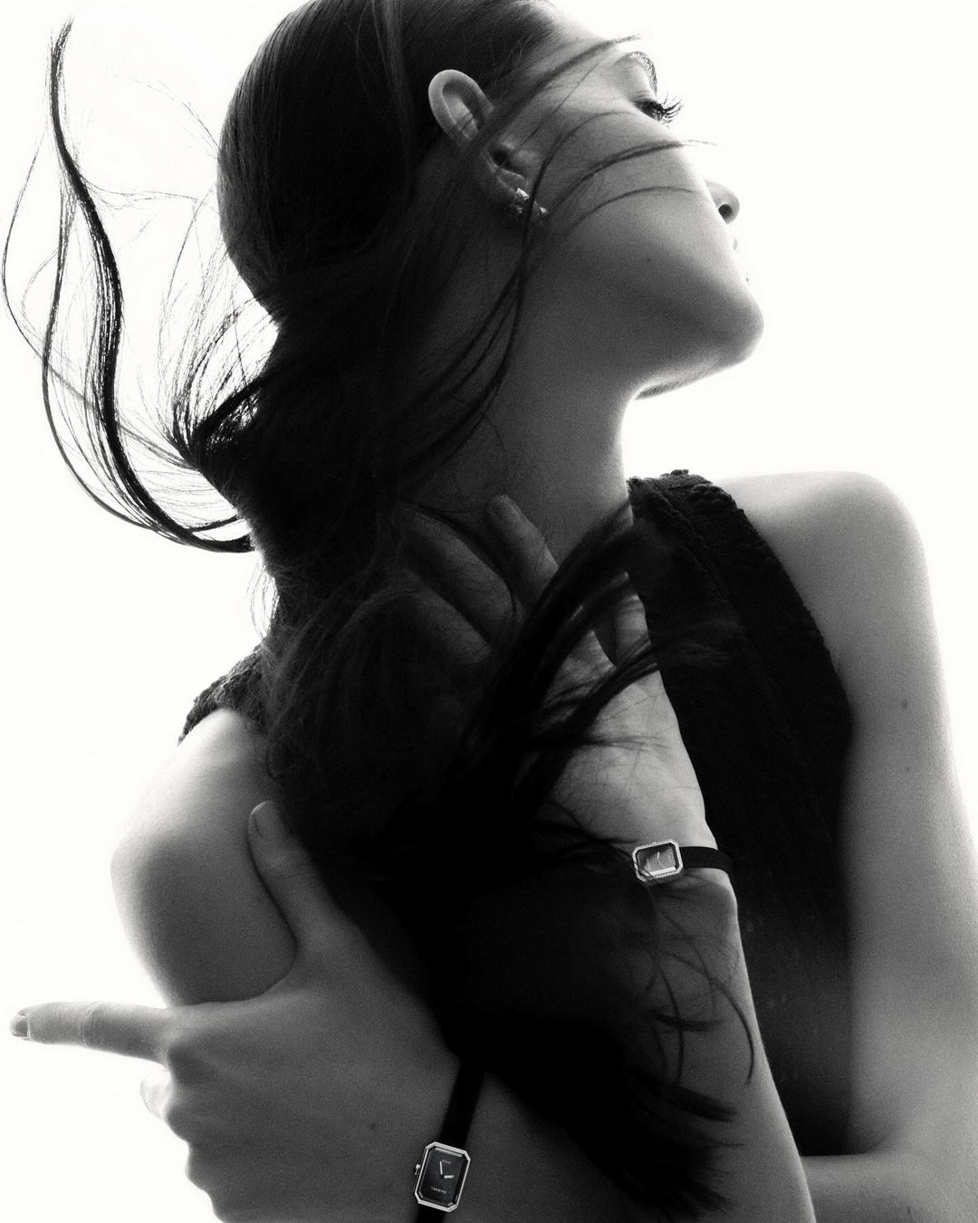 Black and white portrait of a woman with long dark hair, wearing a watch, with her arms crossed and fingers pointing. High-key lighting.