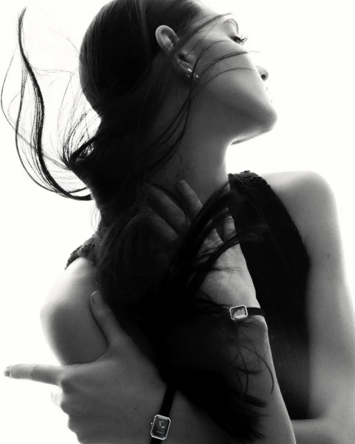 Black and white portrait of a woman with long dark hair, wearing a watch, with her arms crossed and fingers pointing. High-key lighting.
