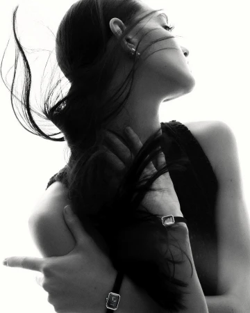 Black and white portrait of a woman with long dark hair, wearing a watch, with her arms crossed and fingers pointing. High-key lighting.