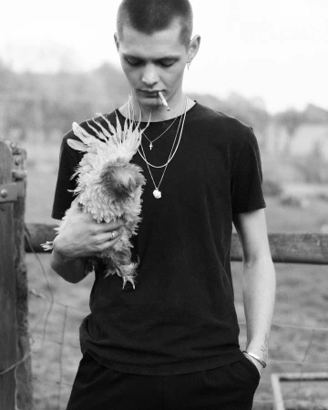 Young man with short hair wearing a black t-shirt holding a fluffy chicken while smoking a cigarette outdoors.