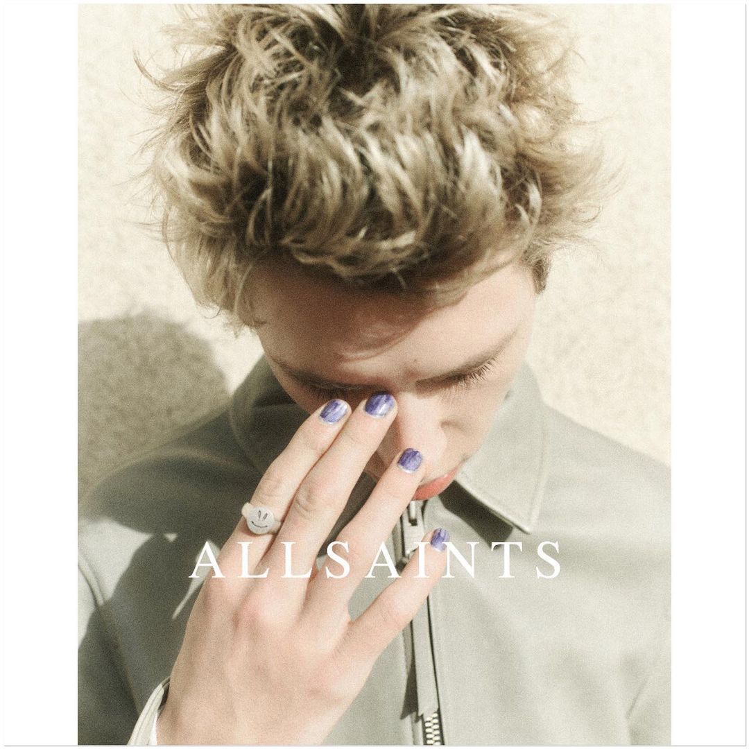 Close-up portrait of a young adult with blond curly hair, wearing a light grey jacket, and blue nail polish by All Saints.