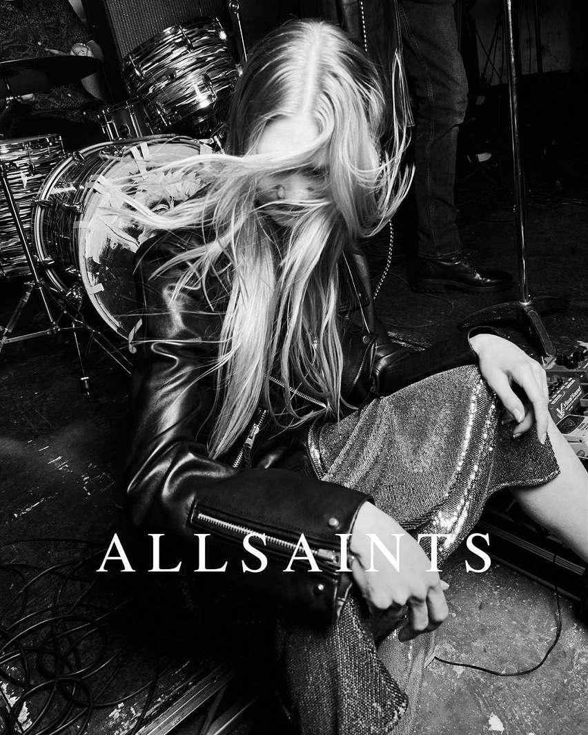Monochrome studio shot: woman with long hair in leather jacket and sequined pants seated near drum set. ALLSAINTS logo visible.