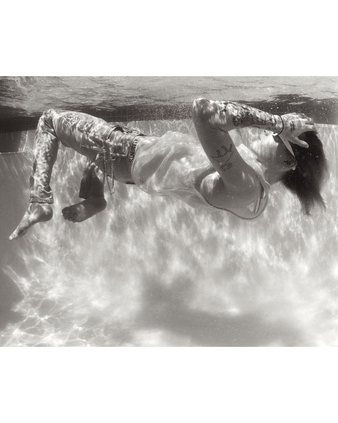 Black and white underwater portrait of a tattooed person in jeans and a white top, with hands behind their head, in swimming pool.
