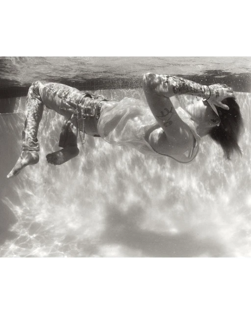 Black and white underwater portrait of a tattooed person in jeans and a white top, with hands behind their head, in swimming pool.