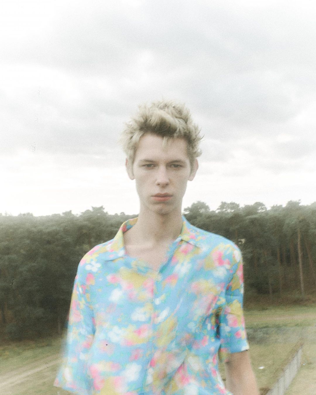 Portrait of a young man with blond hair wearing a floral shirt, posing outdoors with a background of dense forest and overcast sky.