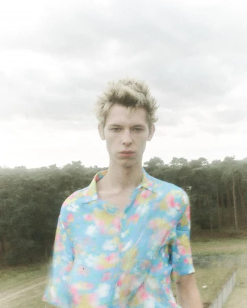 Portrait of a young man with blond hair wearing a floral shirt, posing outdoors with a background of dense forest and overcast sky.