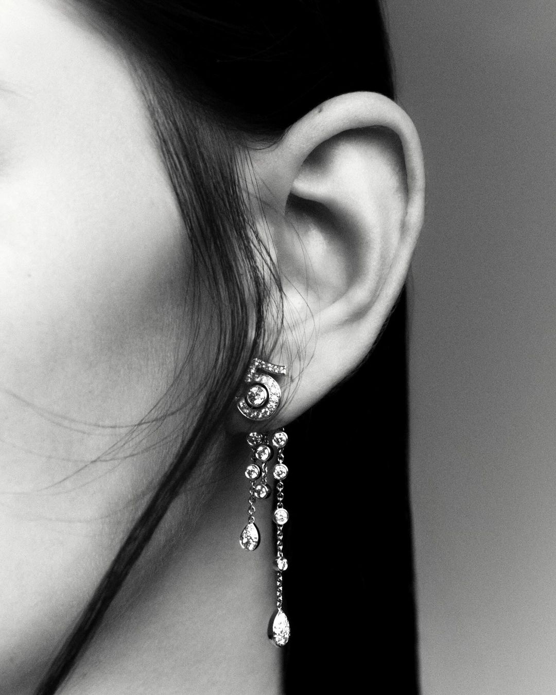 Close-up studio shot of an adult woman's ear wearing diamond 'number 5' earrings with a grey background in monochrome.