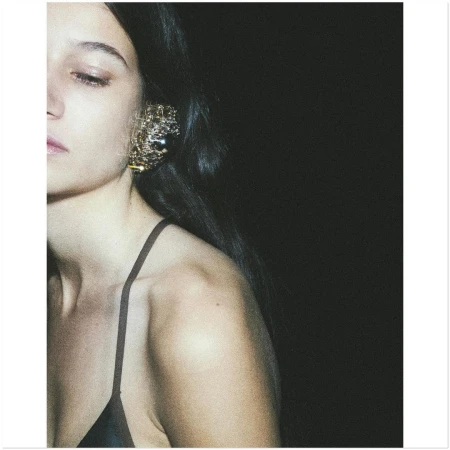 Close-up portrait of a woman wearing a large textured gold ear cuff and dark spaghetti strap top against a dark background.