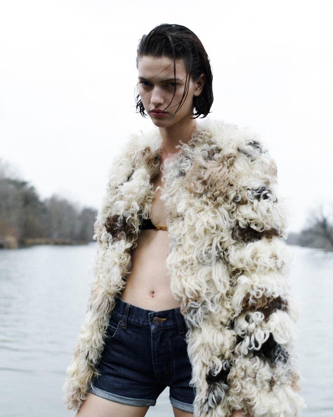 Woman in a shaggy fur coat and denim shorts stands outdoors near a river under an overcast sky, creating a fashion editorial image.