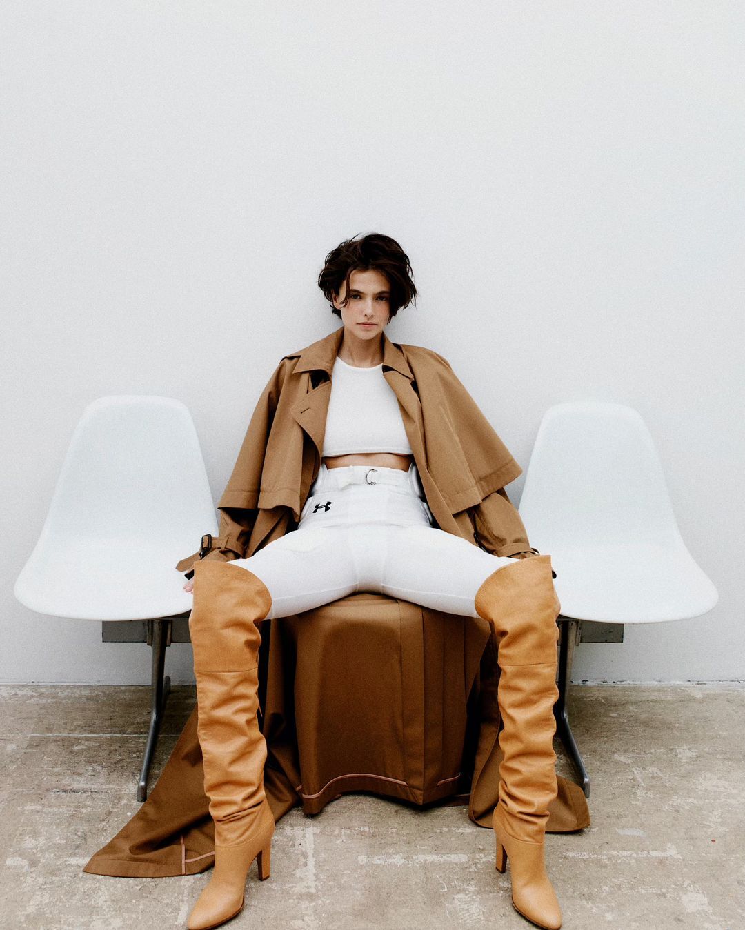 A brunette woman sits between two white chairs wearing a tan trench coat, a white crop top, white pants and tan, high-heeled boots.