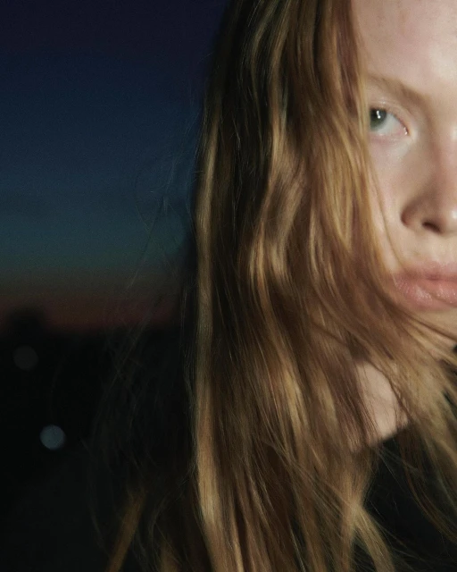 Close-up portrait of a young adult woman with long red hair and blue eyes against a dark background with soft, diffused lighting.