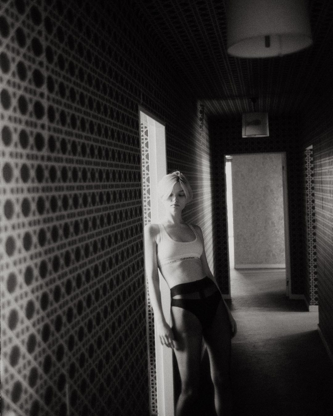 Black and white shot of woman in bra and underwear leaning against patterned wall in hallway. Indoor, low-key lighting.