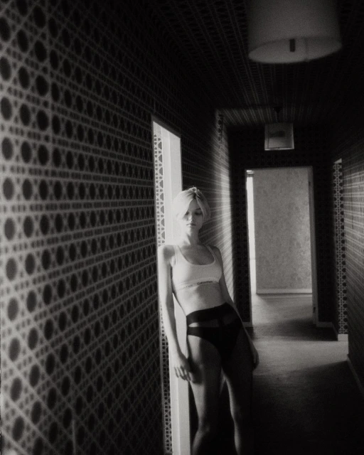 Black and white shot of woman in bra and underwear leaning against patterned wall in hallway. Indoor, low-key lighting.