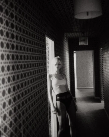 Black and white shot of woman in bra and underwear leaning against patterned wall in hallway. Indoor, low-key lighting.
