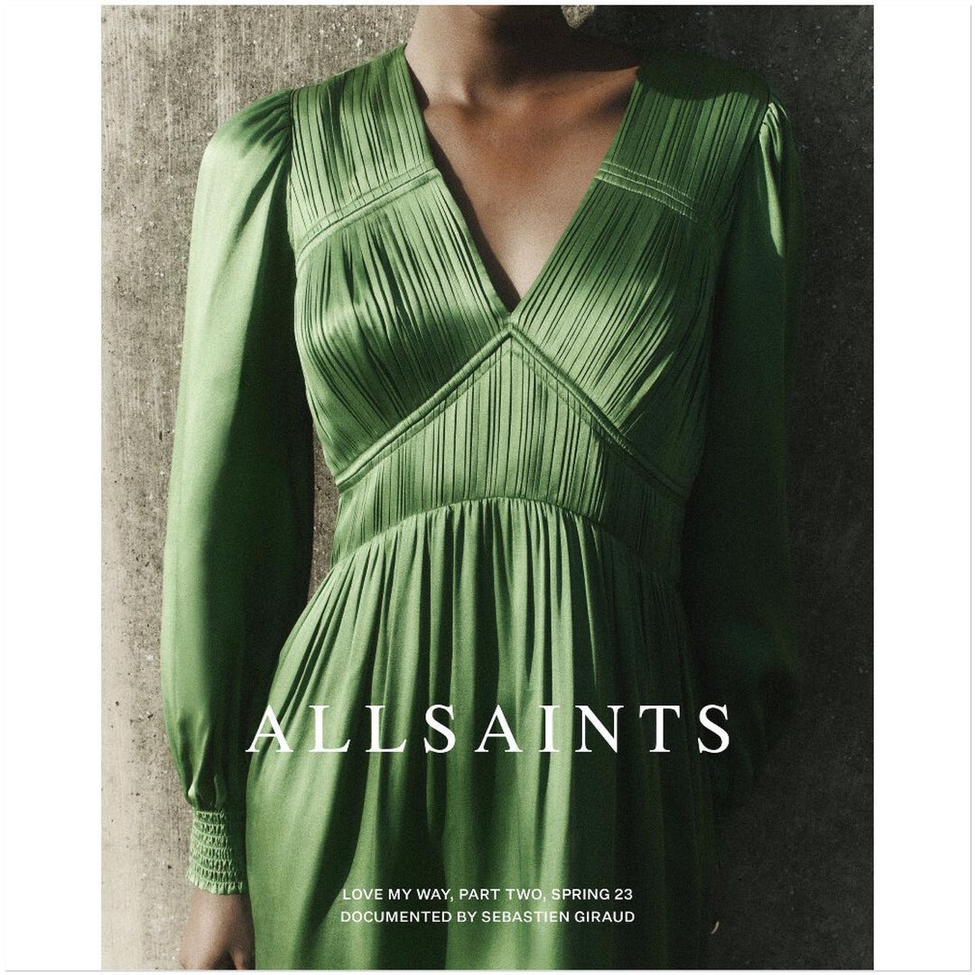 Close-up of a woman in a pleated green satin dress from AllSaints' Spring 23 collection, indoors against a textured grey wall.