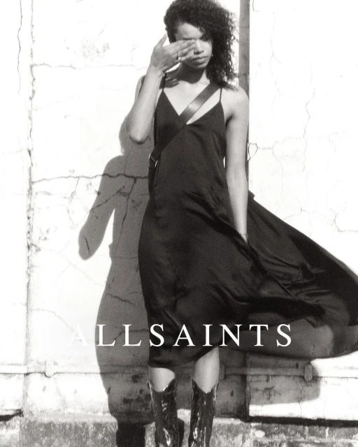 A woman in a slip dress and boots stands in front of a textured wall, styled for a fashion advertisement in black and white.