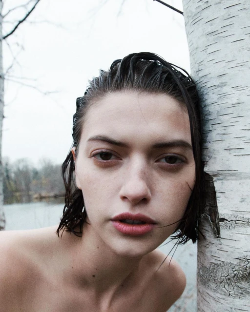 Close-up portrait of a young adult woman with wet dark hair, fair skin, next to a birch tree, set against an overcast sky and lake.