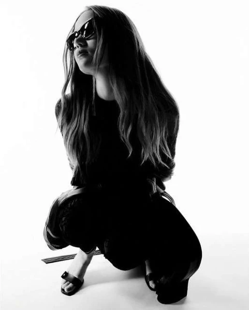 Monochrome portrait of a young woman squatting and wearing sunglasses, set against a bright white backdrop in a studio setting.