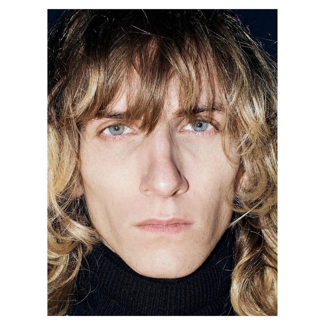 Portrait of a blonde adult wearing a black turtleneck sweater, against a dark background. The person has fair skin and light-colored eyes.