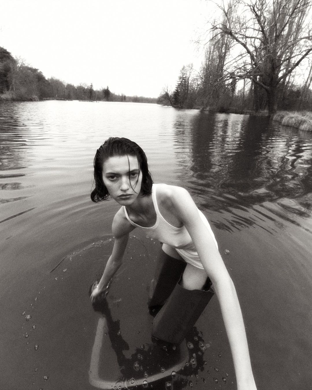 Monochrome portrait of a solitary woman in a white tank top and tall boots standing in a lake with trees and sky in the background.