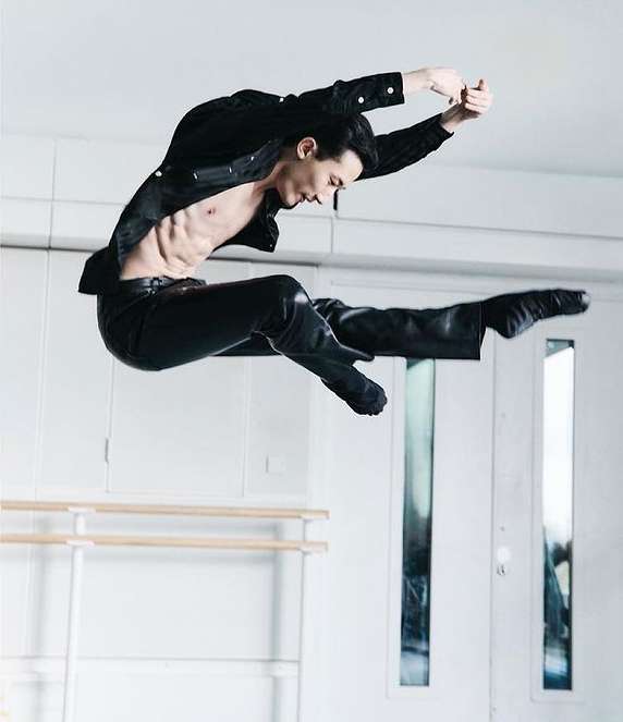 An athletic adult male in a black outfit jumps mid-air against a bright white indoor setting. He is shirtless, with legs split, arms raised.