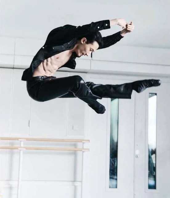 An athletic adult male in a black outfit jumps mid-air against a bright white indoor setting. He is shirtless, with legs split, arms raised.