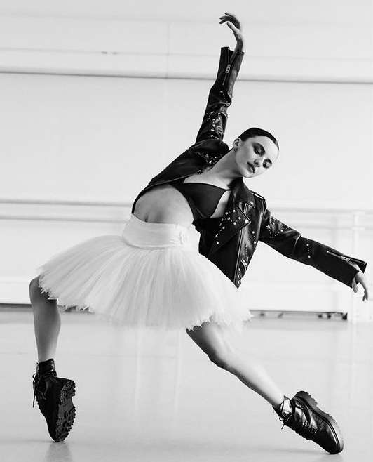 An adult female ballerina in a tutu and leather jacket strikes a pose indoors, backlit in a dance studio against a white wall.