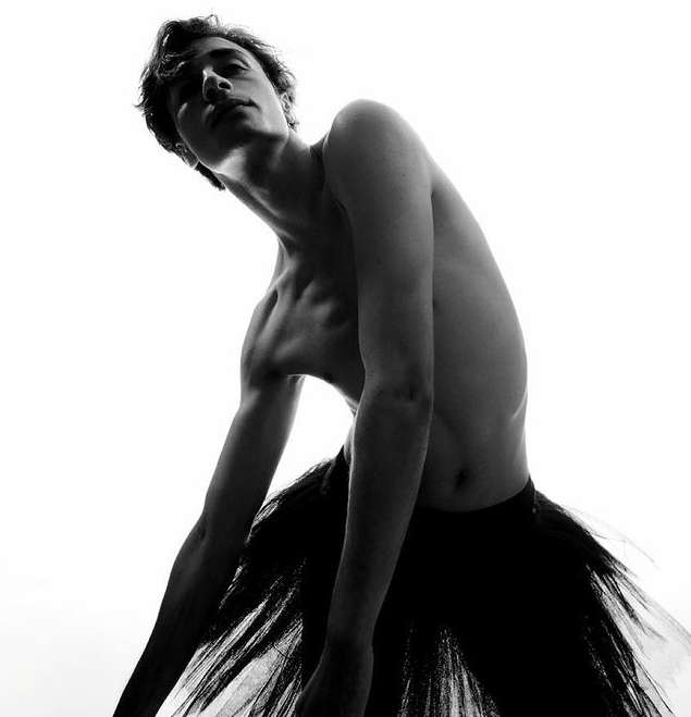 Black and white shot of a lean male dancer in a tutu, looking up with a serious expression, posing against a bright white backdrop.