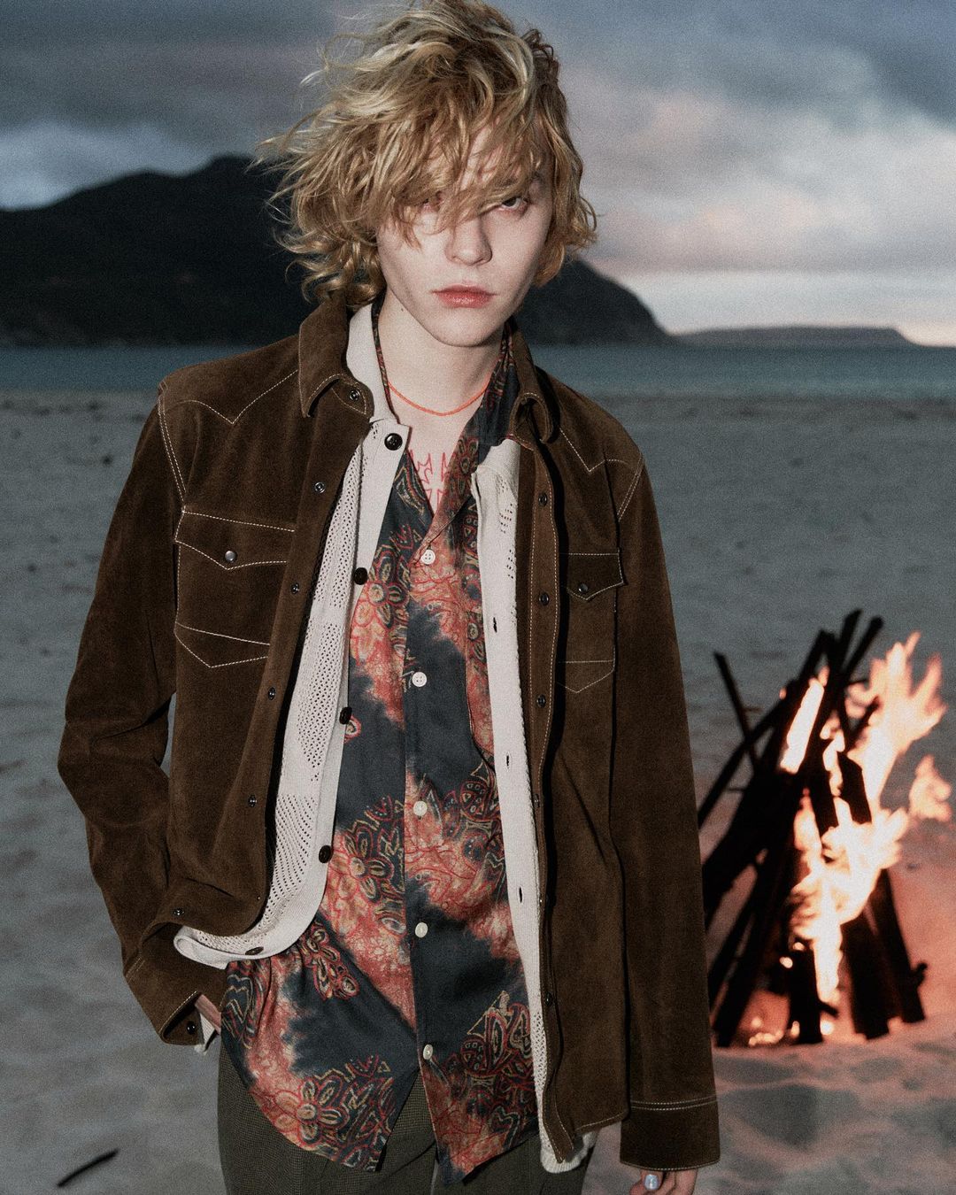 A blonde adult male stands on a beach in front of a campfire and mountain backdrop, wearing a brown suede jacket and patterned shirt.
