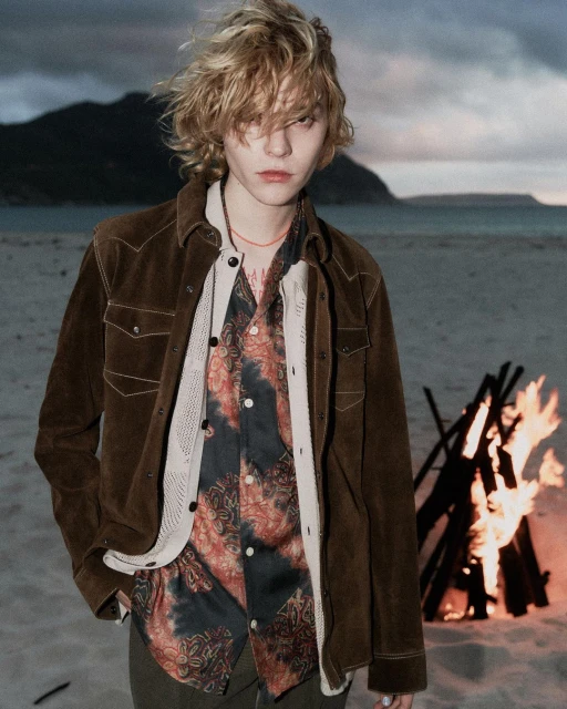A blonde adult male stands on a beach in front of a campfire and mountain backdrop, wearing a brown suede jacket and patterned shirt.