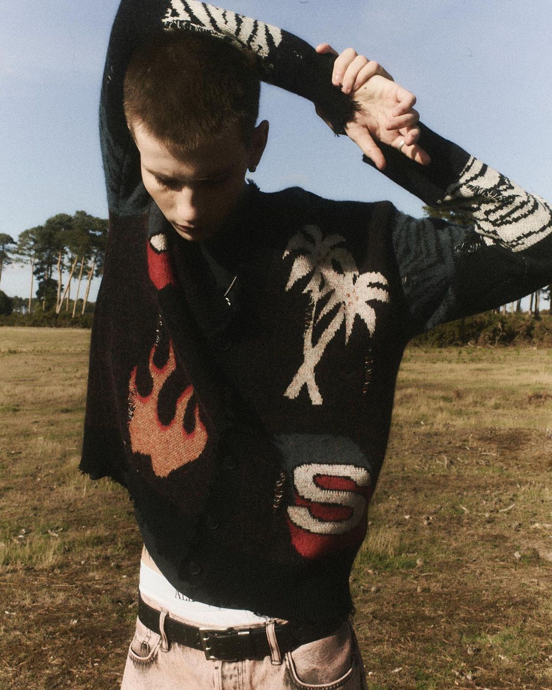A slender young man with short hair stands in a field wearing a black patterned sweater with pink jeans and a black belt, looking down.