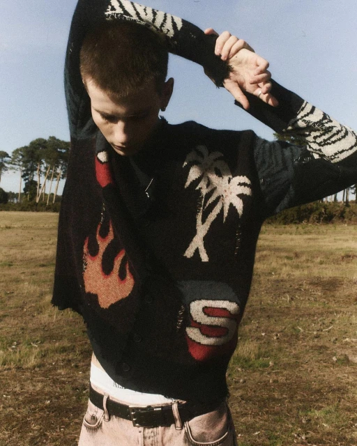 A slender young man with short hair stands in a field wearing a black patterned sweater with pink jeans and a black belt, looking down.