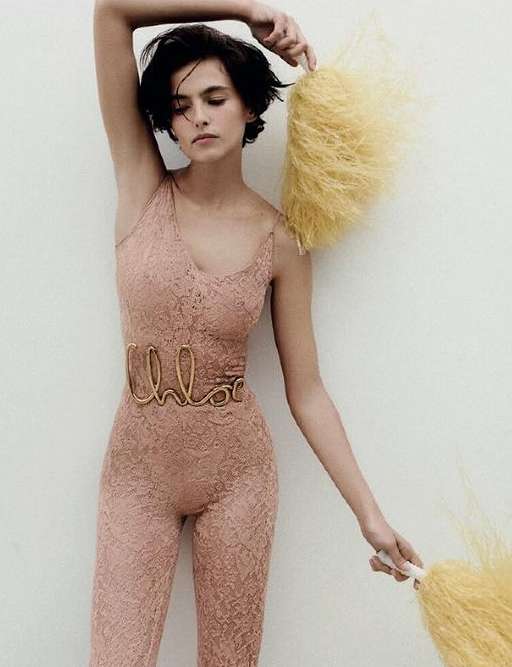 A woman in a lace catsuit strikes a pose against a plain backdrop, holding feathered fans in a fashion-forward editorial shot.