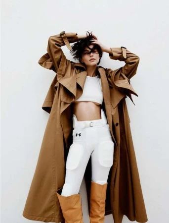 Woman in a white crop top, football pants, and boots, striking a pose with hands on her head in front of a plain white wall.
