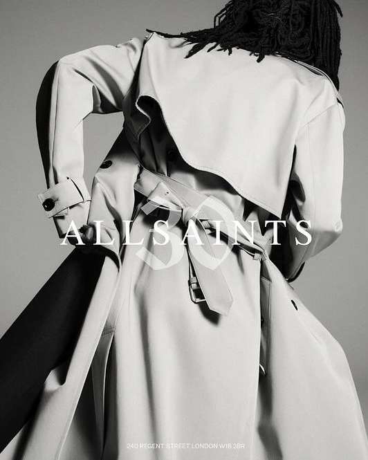 Back view of person in light-colored trench coat with belt, and dark dreadlocks. The brand name ALLSAINTS is overlaid on the image.