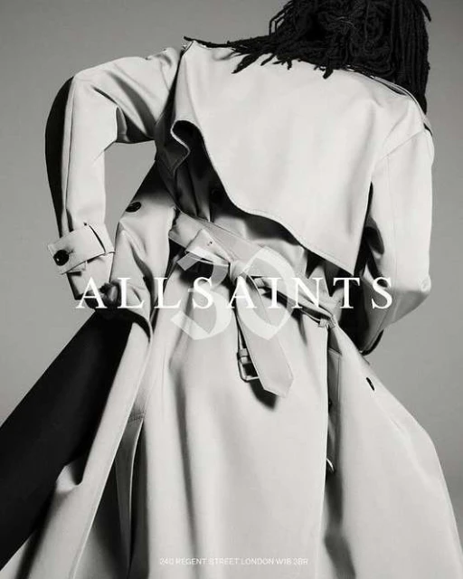 Back view of person in light-colored trench coat with belt, and dark dreadlocks. The brand name ALLSAINTS is overlaid on the image.