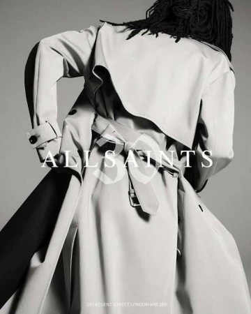 Back view of person in light-colored trench coat with belt, and dark dreadlocks. The brand name ALLSAINTS is overlaid on the image.