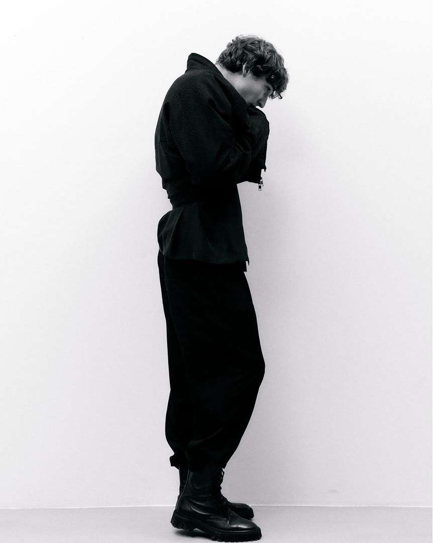 Black and white full-body shot of a person in a dark jacket, baggy pants, and combat boots, standing against a light-colored wall.