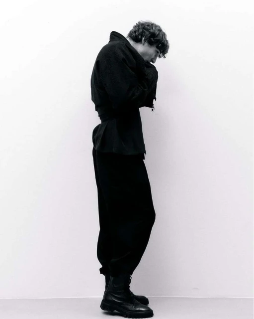 Black and white full-body shot of a person in a dark jacket, baggy pants, and combat boots, standing against a light-colored wall.