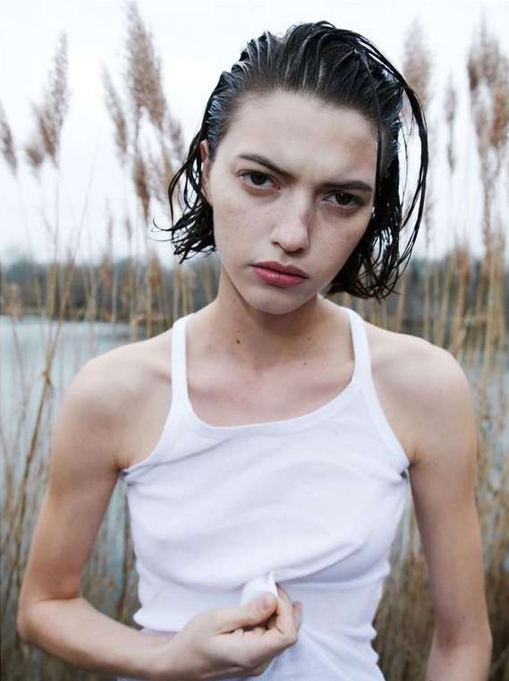 Wet-haired model wearing a white tank top outdoors against tall dry grass by water, photographed by Sebastien Giraud