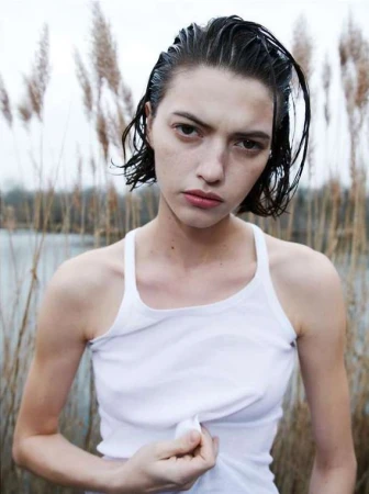 Wet-haired model wearing a white tank top outdoors against tall dry grass by water, photographed by Sebastien Giraud