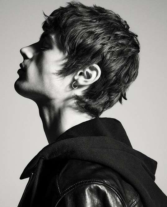 Profile portrait of a young man with dark textured hair and hoop earring wearing a black jacket against a plain grey background
