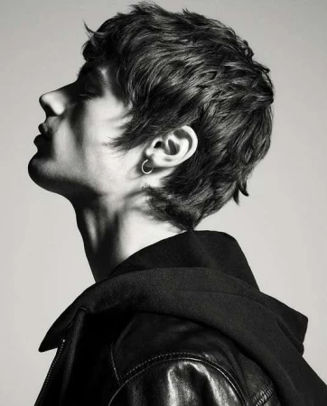 Profile portrait of a young man with dark textured hair and hoop earring wearing a black jacket against a plain grey background