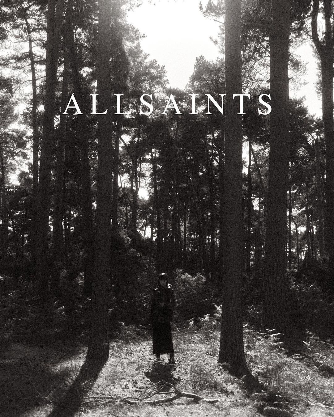 Monochrome photograph features a single female figure in a hat and coat standing in a dense pine forest in full-length shot.