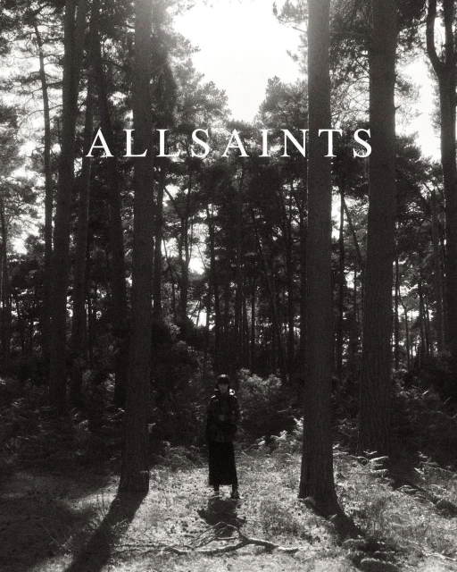 Monochrome photograph features a single female figure in a hat and coat standing in a dense pine forest in full-length shot.